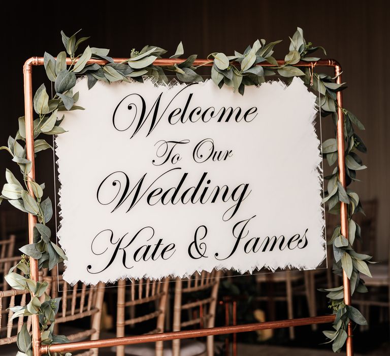 White acrylic wedding welcome sign on copper frame surrounded by eucalyptus 