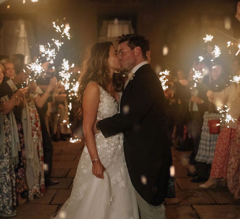 Bride in floral Mira Zwillinger wedding dress kissing groom as they stand inbetween their wedding guests for their sparkler send off 