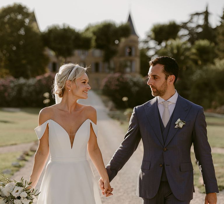 Bride wears pearl bridal headband and carries white rose bouquet as she holds her grooms hand who wears three piece blue suit 