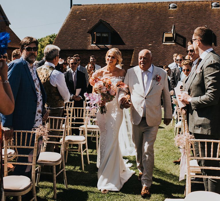 Father of the bride in a light suits walks the bride in an off the shoulder wedding dress down the aisle 