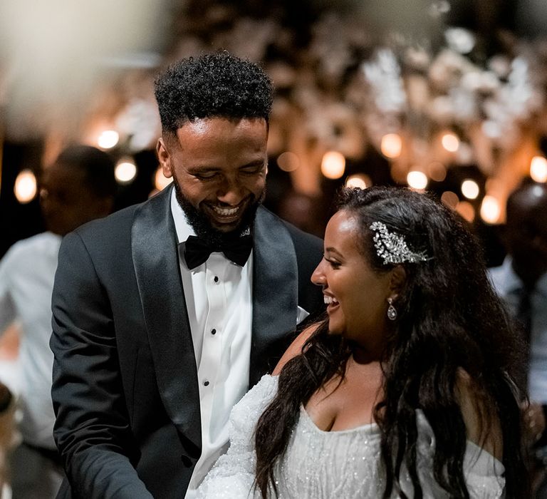 Bride wears long-sleeve embellished reception dress whilst stood beside her groom in black tie as they cut their wedding cake