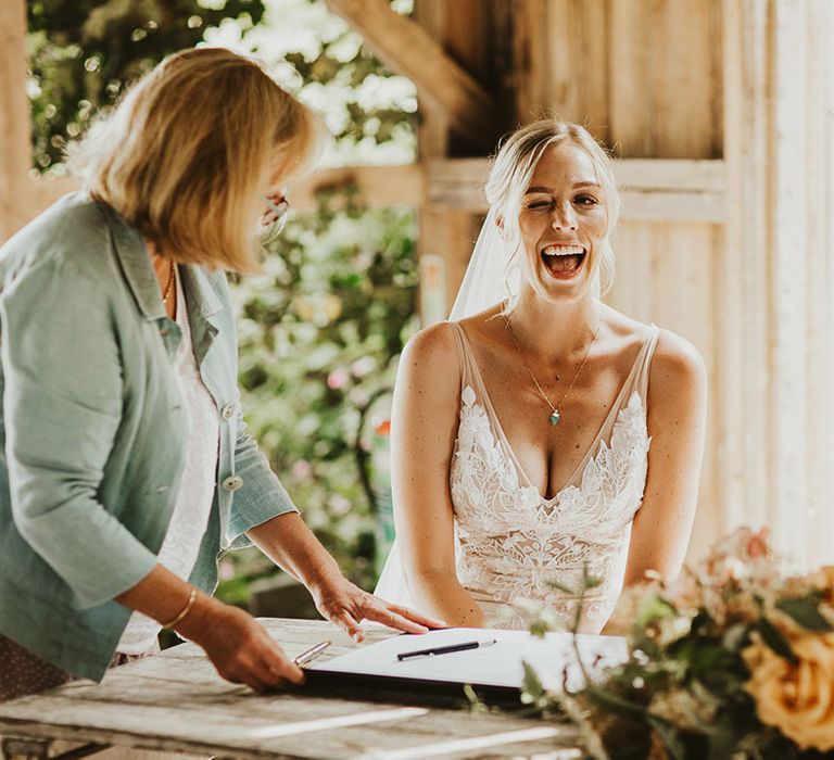 Bride gives a cheeky wink as she wears a lace Made With Love wedding dress and signs the register with the groom in a light suit 