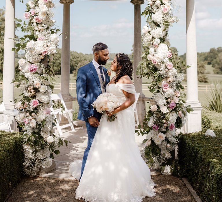 Bride & groom stand beneath wedding dome complete with white and pink pastel florals with green foliage