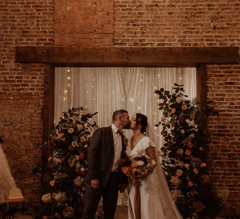 Pink rose flower columns with white wedding drapes and fairy lights decorate the altar with the bride in a Emma Beaumont Atelier dress and groom in a custom suit