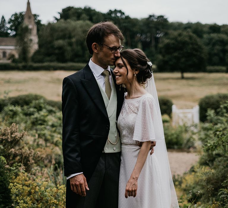 Groom in three piece suit with sage green waistcoat and tie kissing bride in batwing sleeve lace detail wedding dress on the head