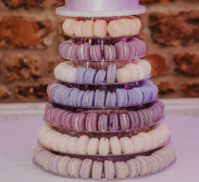 Single tier white iced wedding cake with a purple ribbon on top of a display of lilac, purple, lavender and violet macarons 