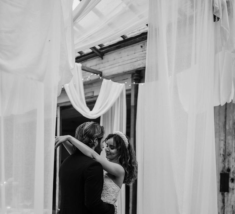 Bride in sleeveless lace wedding dress with pearl details, pearl headband and silver belt dancing with groom in black tux by ceiling drapes decoration