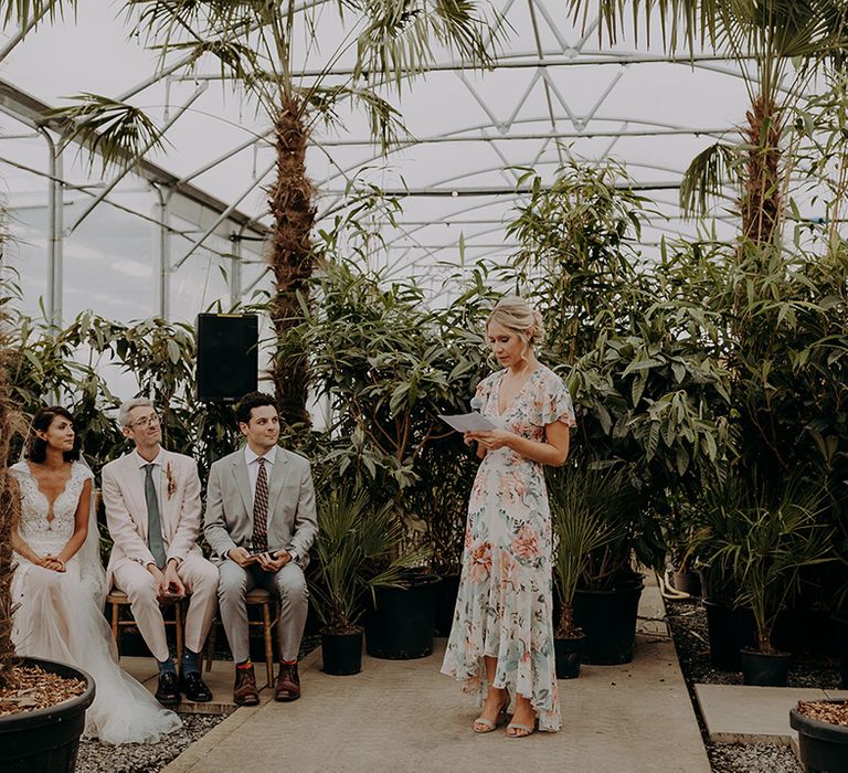 Bridesmaid in floral dress does reading at front of ceremony with greenery in background