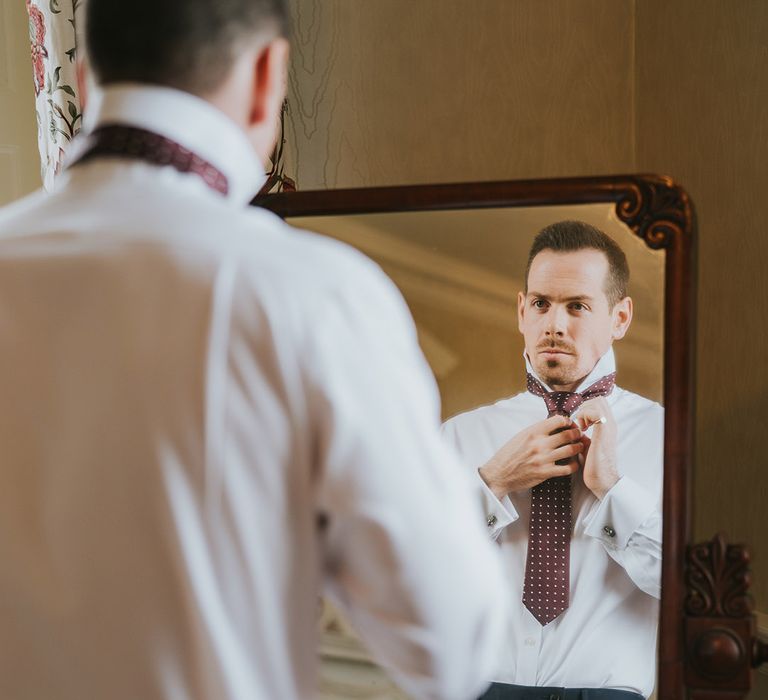 Groom puts on his burgundy tie with white spots as he prepares for the wedding 