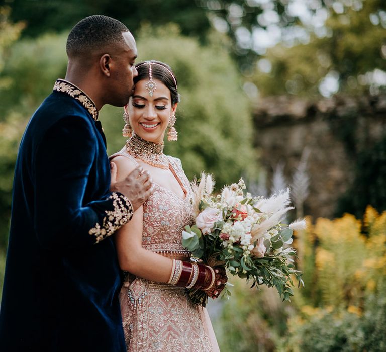 Groom wearing midnight blue and gold Indian wedding outfit kissed bride in golden bridal Lehnga