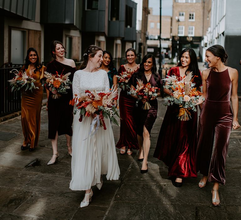 Bride in long vintage wedding dress walking with bridesmaids in mismatched velvet dresses down the streets of London, all holding large autumnal bouquets with orange, black, green and yellow roses, peonies, foliage and pampas grass