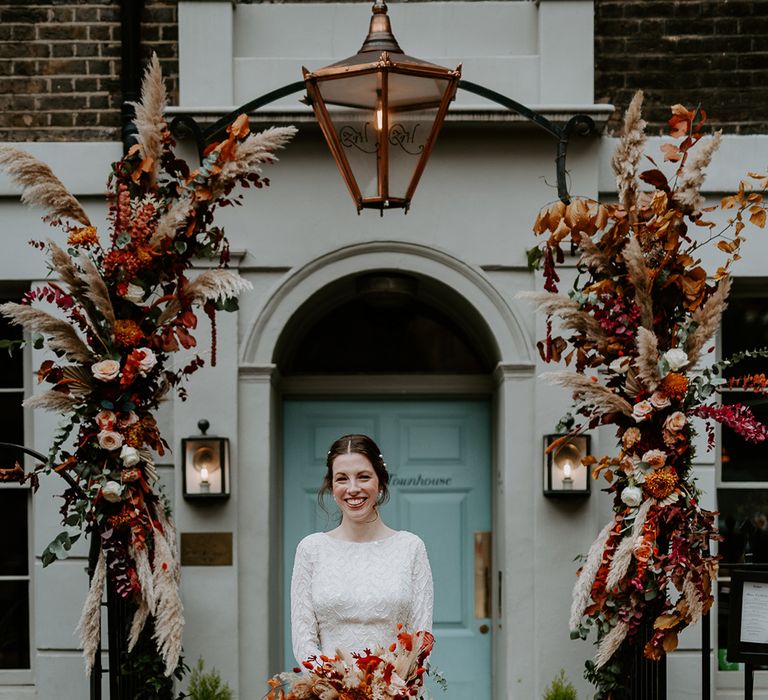 Bride in long sleeve lace vintage wedding dress and closed toe satin heels holding large autumnal bouquet and standing at The Zetter Townhouse with large floral arch with roses, peonies, foliage and pampas grass