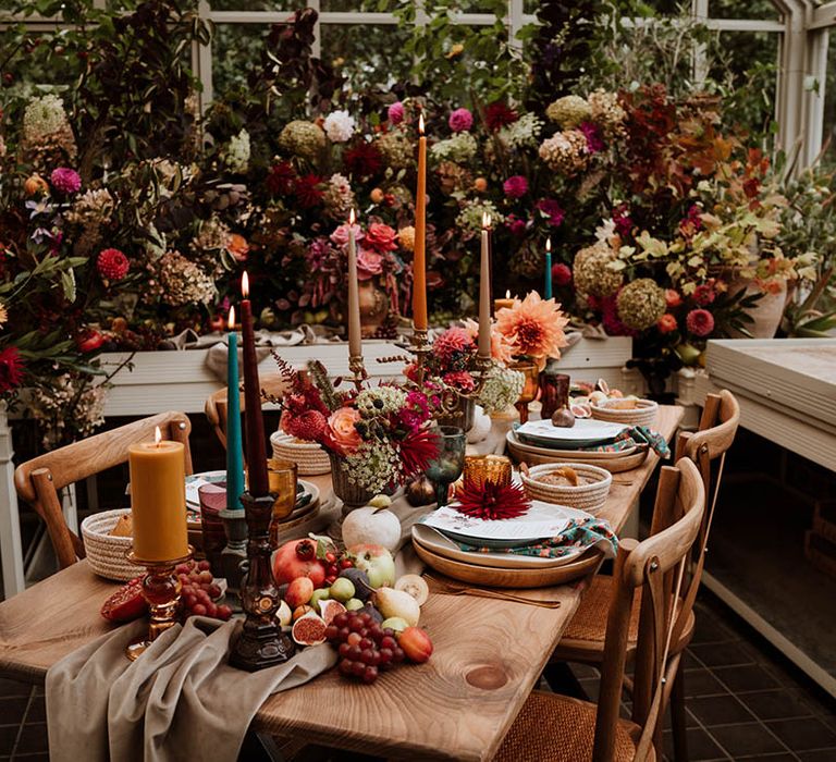 Autumnal wedding table plan with neutral-toned tablerunner on rustic wooden table, orange, green and red coloured glasses, earthenware, seasonal fruit and vegetables, red, brown and green tapered candles in gold candlesticks and clear vases with autumnal coloured flowers