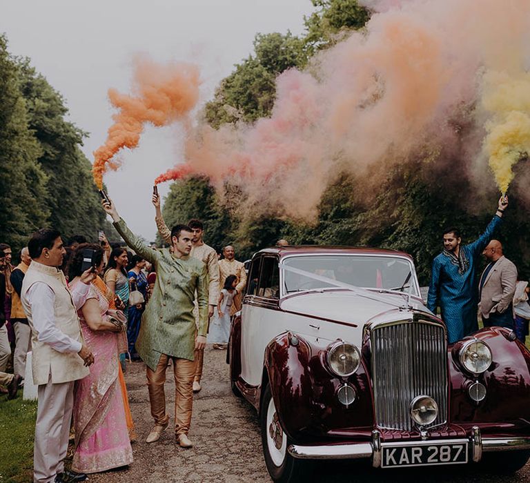 Wedding guests carry colourful smoke bombs as they walk alongside vintage wedding car 