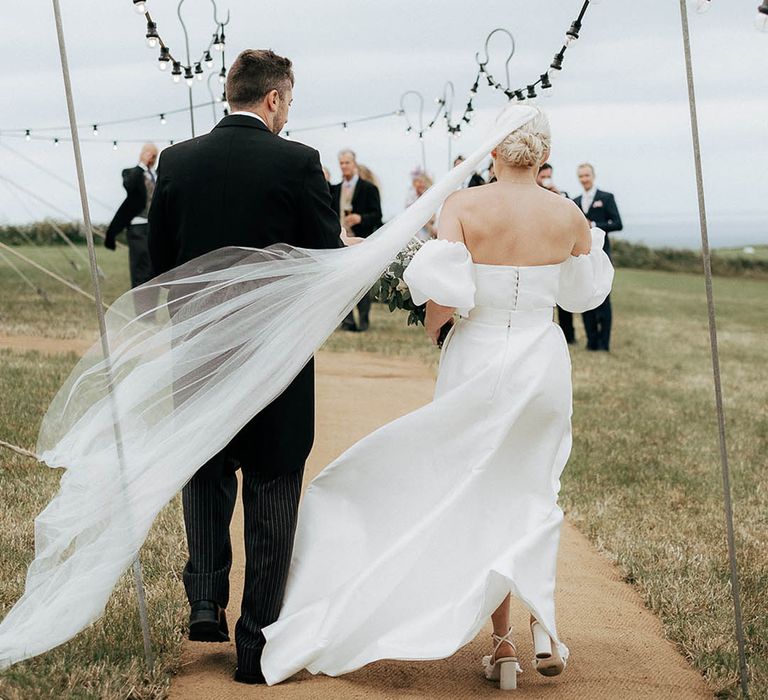 Bride wears floor-length veil in her blonde hair as she walks alongside her groom toward marquee reception 