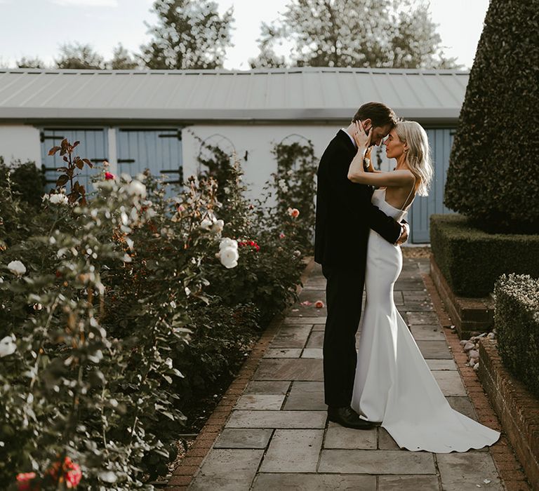 Groom in traditional black tuxedo stands embracing the bride in a fitted wedding dress with thin spaghetti straps posing for their couple portraits 