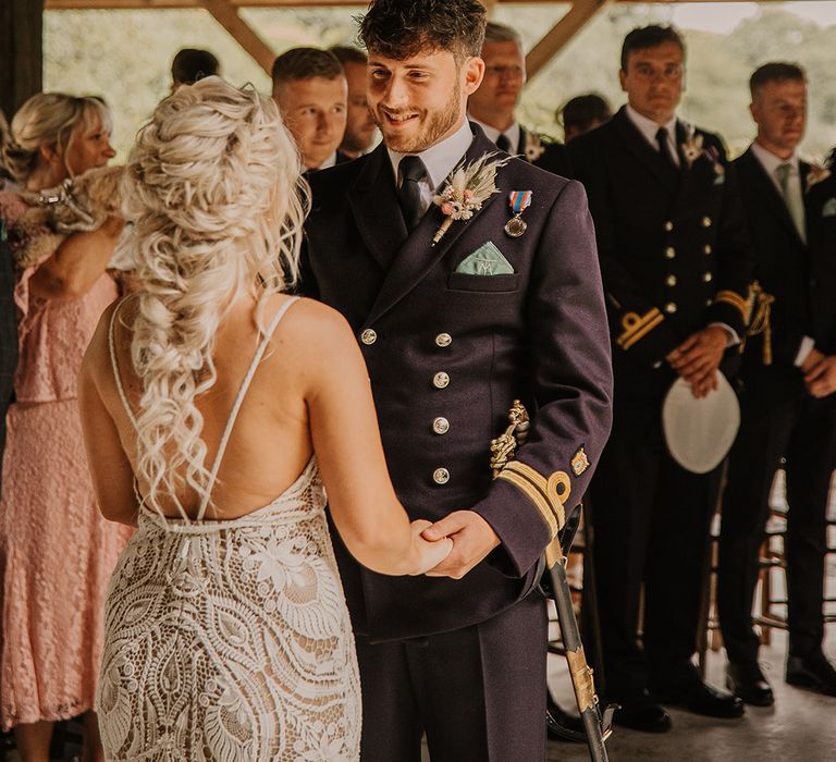 The groom in a double breasted military uniform with a pampas grass buttonhole smiling at the bride as they stand for their ceremony 