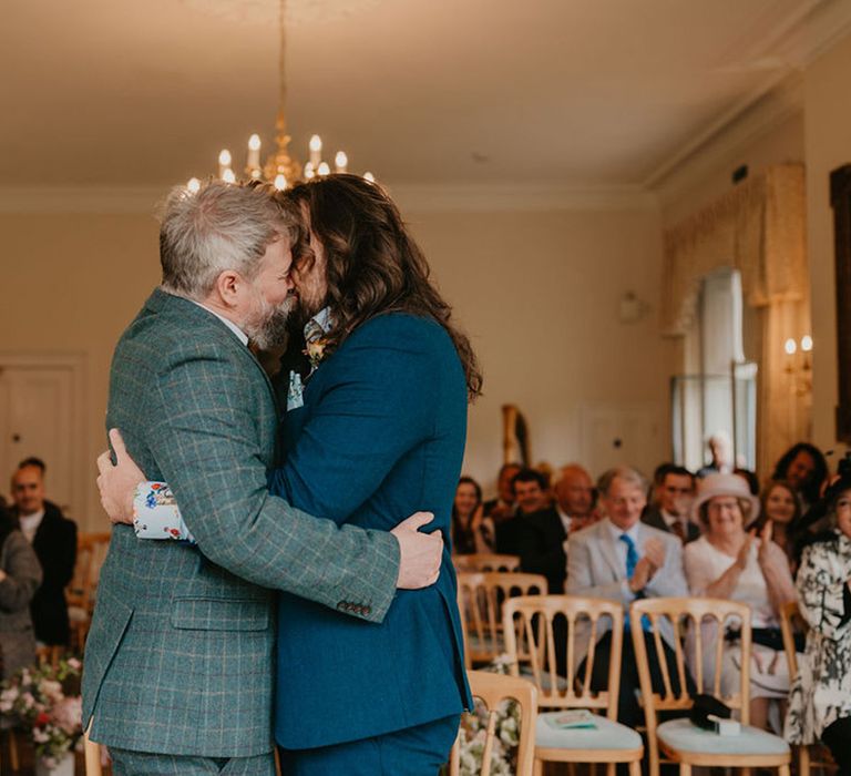 Groom in tartan suit and groom in blue suit kiss during wedding ceremony at the Cambridge Cottage in Kew Gardens 