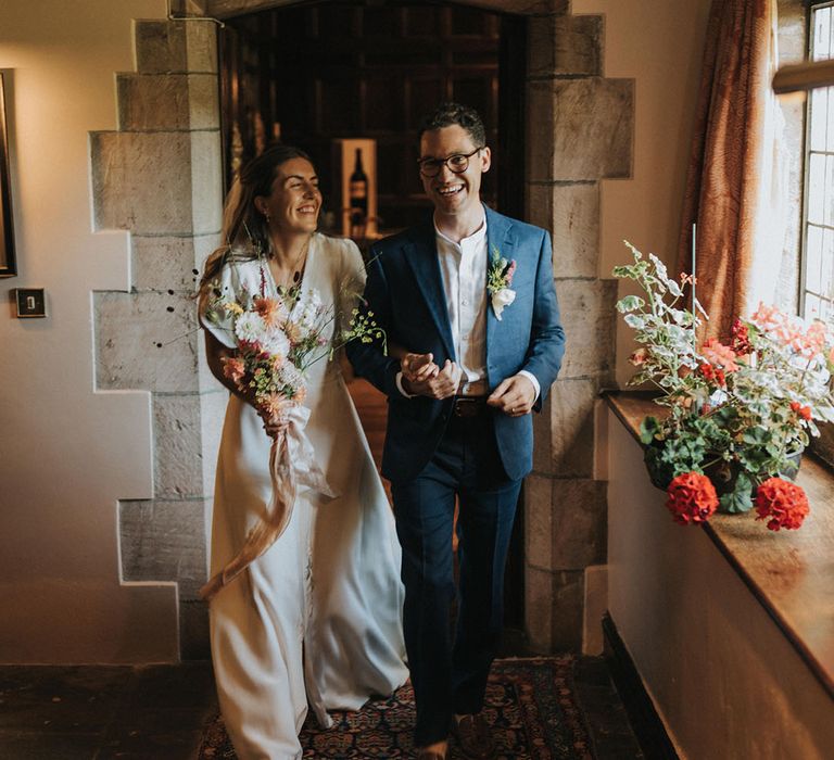 Groom in navy suit and white shirt and glasses walking with the bride in a puff sleeve wedding dress with rustic wedding flowers bouquet and buttonhole 