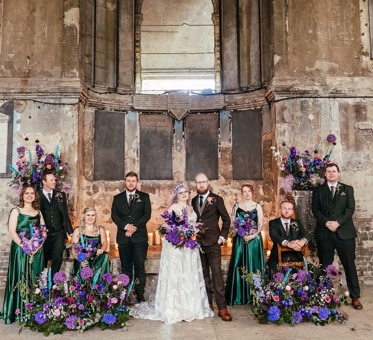 Bride in appliqué sheer Evie Young bridal gown and cathedral length veil standing with groom in mocha brown textured suit with deep green tie and pink and purple boutonniere, holding bright purple bridal bouquet with purple hydrangeas, pink garden roses, purple orchids, lilac carnations, dried flowers and peacock blue feathers standing with bridesmaids in tie strap forest green bridesmaid dresses and groomsmen in dark three piece textured suits with dried flower boutonnieres, surrounded by large purple, blue and pink floral displays 