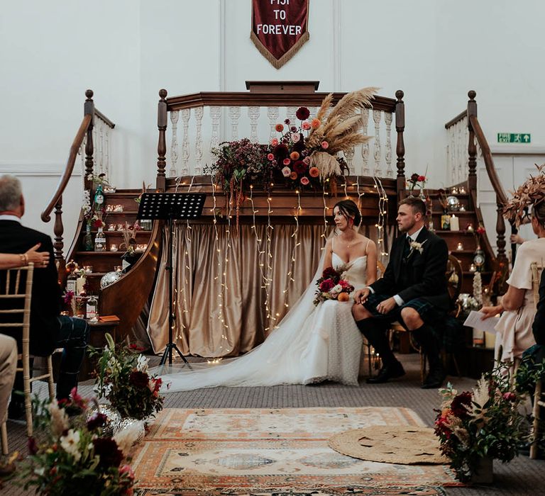 boho wedding ceremony decor with rugs, fairy lights, pampas grass, candles, wildflowers and a fabric banner 