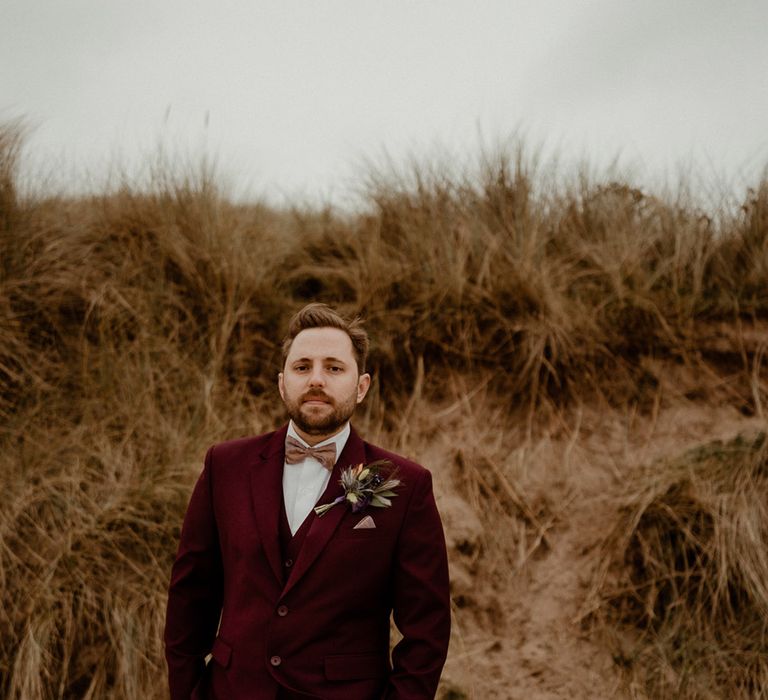 Groom in three piece burgundy suit with a dried flower peacock feather buttonhole on the beach at Newton Hall 