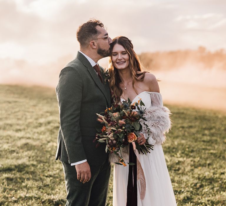 Groom in three piece suit with dark orange brown tie smiling with the bride in an off the shoulder wedding dress with stunning views of the Cotswolds