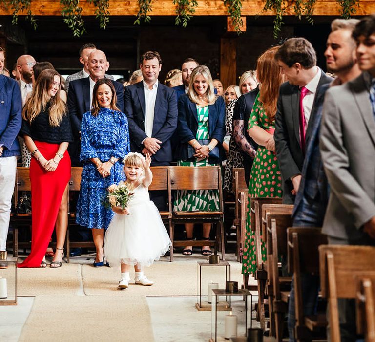 Flower girl in white tulle dress and headband walks down the aisle holding a small bouquet 