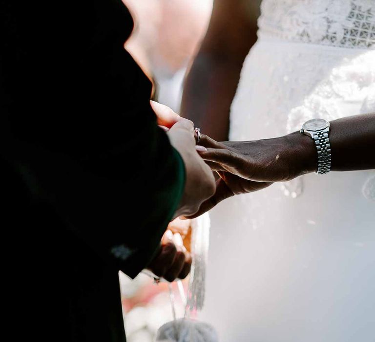 Bride in v neck sleeveless lace Pronovias wedding dress and silver Rolex watch exchanging rings with groom in velvet grooms blazer 