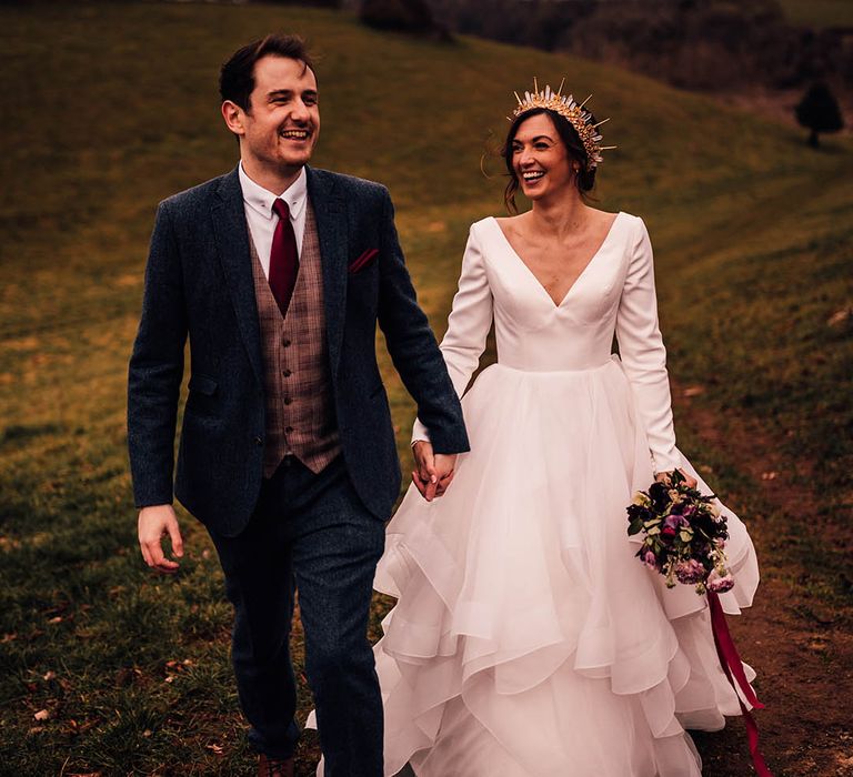 Bride in long sleeve Morilee wedding dress and groom in navy three piece suit walking hand in hand for wedding 