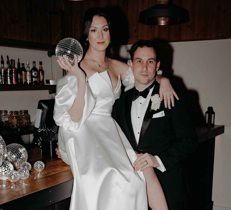 Bride in white silk wedding dress with bespoke puff sleeves and groom in black tuxedo with disco ball decor at Upton Barn wedding venue bar