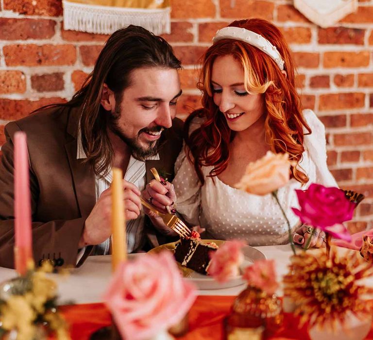 Groom in brown grooms blazer and white shirt eating wedding cake with bride in three quarter length puff sheer sleeve wedding dress with delicate polka dot overlay and square neck and white 3D applique polka dot headband at retro wedding tablescape with pink tapered candles, orange table runner and mismatched single flower vases