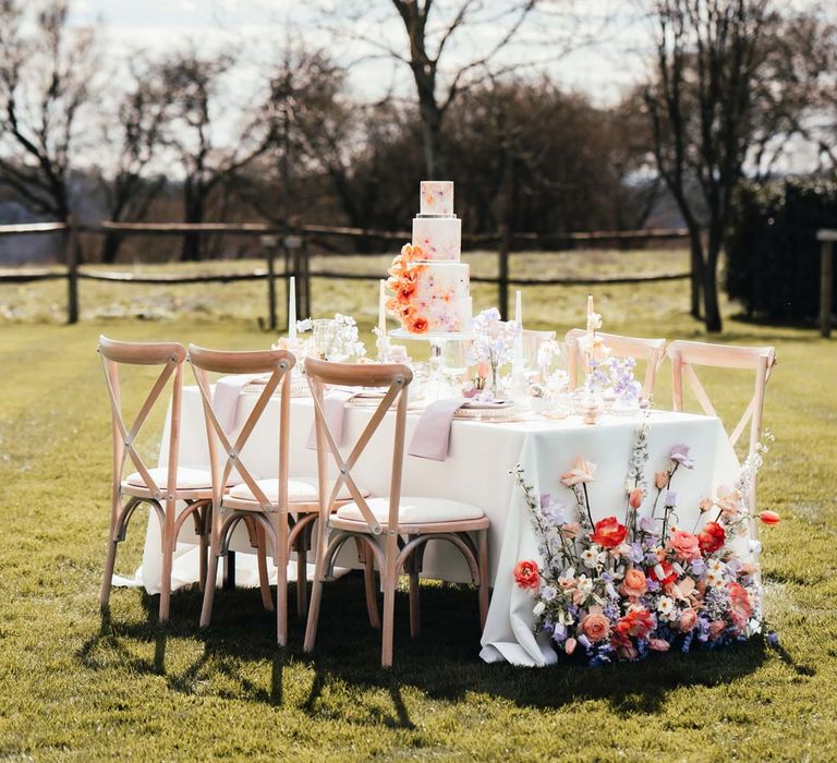 Peach and lilac wedding tablescape with spring flower centrepieces, lilac napkins, tinted glassware and gold cutlery and crockery on white tablecloth at Rackleys Barn with wedding tablescape flower arrangement with peach garden roses, white peonies, lilac sweet peas, bluebells and wildflowers