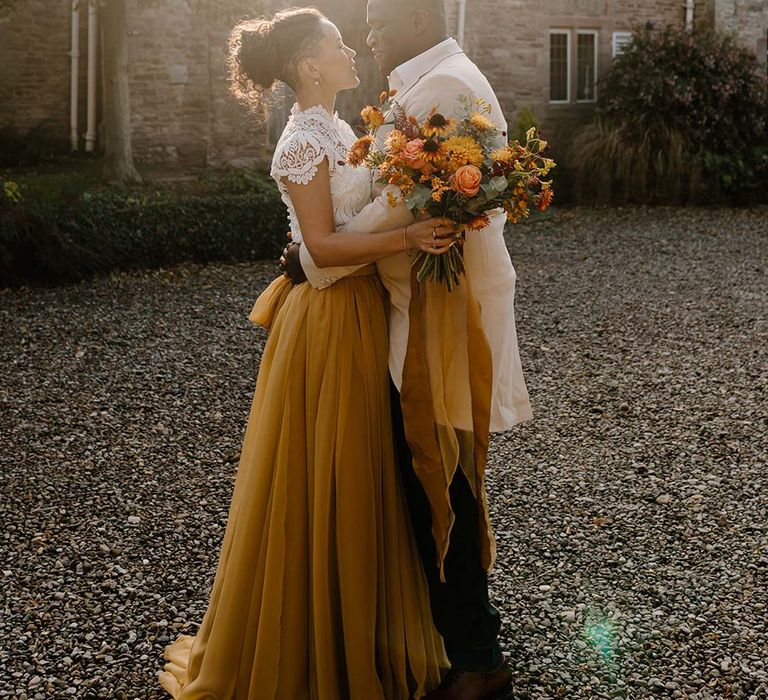 Bride in white mesh lace bridal crop top and yellow wedding skirt holding dried autumnal wedding bouquet with sunflowers, garden roses, pampas grass, eucalyptus, yellow tall kangaroo paws, orange osmanthus flowers, foliage and dried flowers embracing groom in cream grooms morning suit outside of Broadfield Court wedding venue