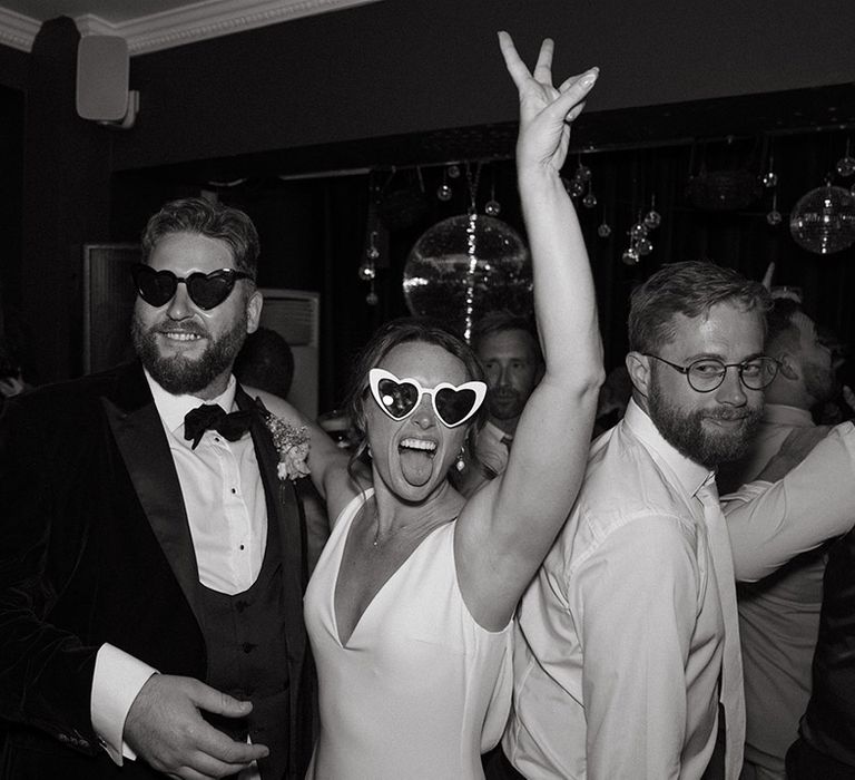 Bride wearing white heart retro wedding sunglasses dancing with the groom in a black tuxedo at the wedding reception 