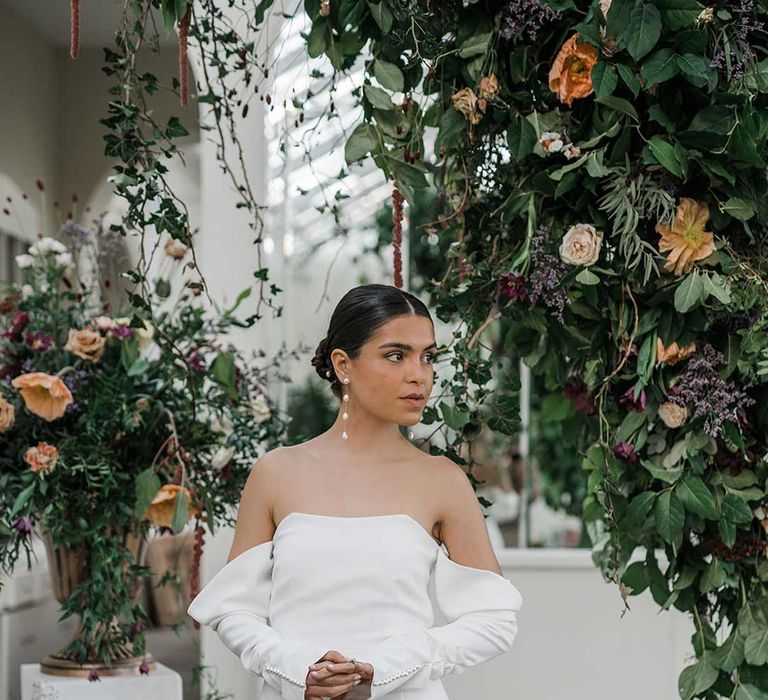 Bride in long sleeve off the shoulder wedding dress with pearl button detailing and a side slit surrounded by suspended wildflower and foliage arrangements in large gold vases at Buxted Park