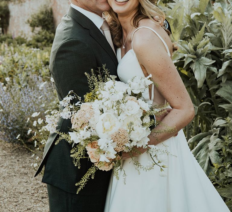 Groom in black suit jacket embracing the bride holding pale pink and white bouquet 