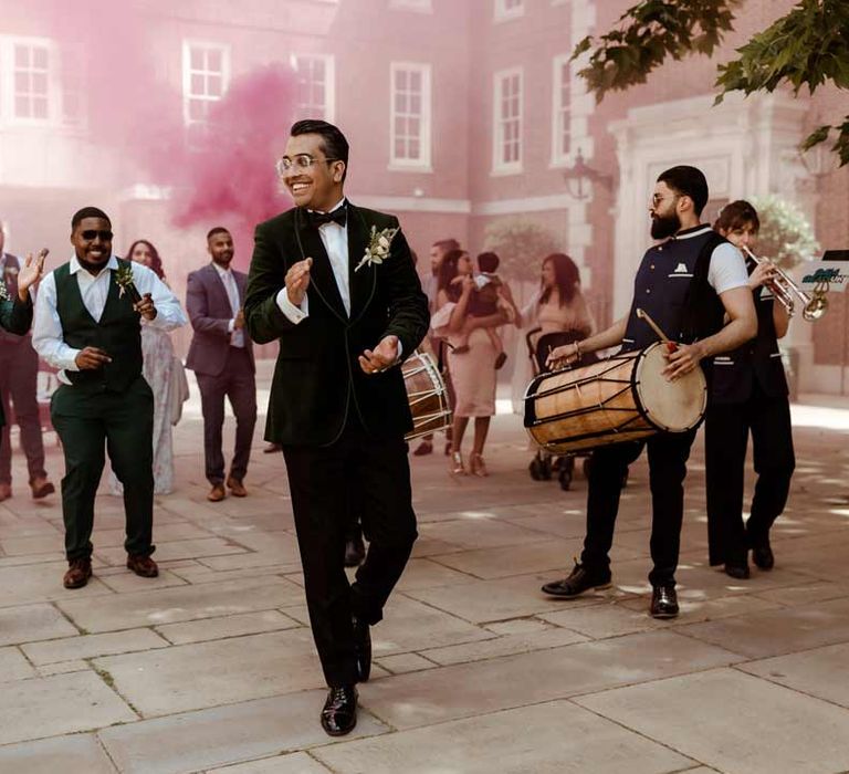 Groom in bottle green velvet grooms suit, black bowtie and chic boutonniere walking through the Inner Temple Hall courtyard with pink smoke bomb in the background