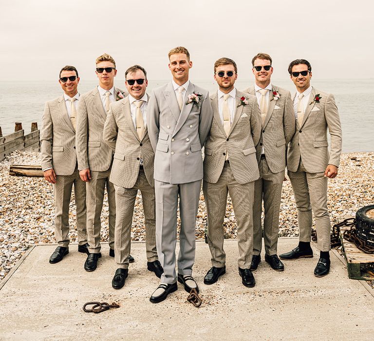 groom in a light grey double breasted jacket standing with his groomsmen in beige suits at beach wedding 