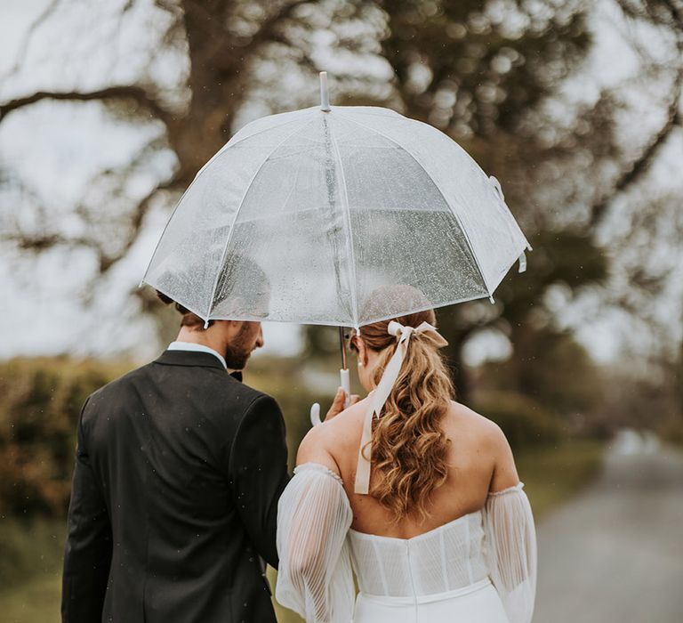 Groom holding clear umbrella walking with bride at Eden Barn rustic wedding with pink hair ribbon