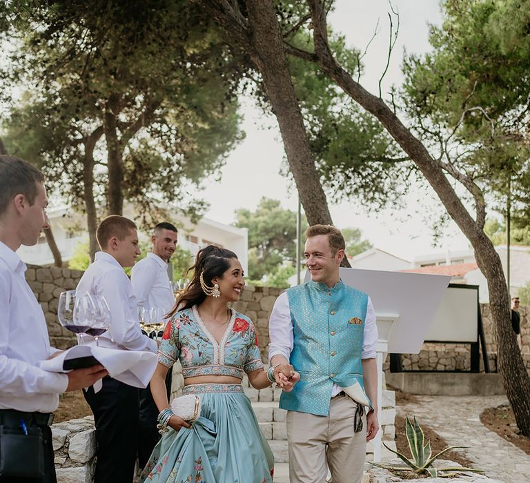 Bride and groom in turquoise traditional south asian wedding outfits walk down steps at wedding welcome party in Hvar Croatia