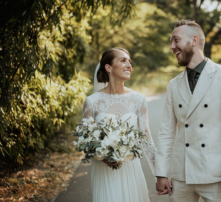 Bride in a Catherine Deane wedding dress with lace long sleeves holding hands with her groom in a beige wedding suite 