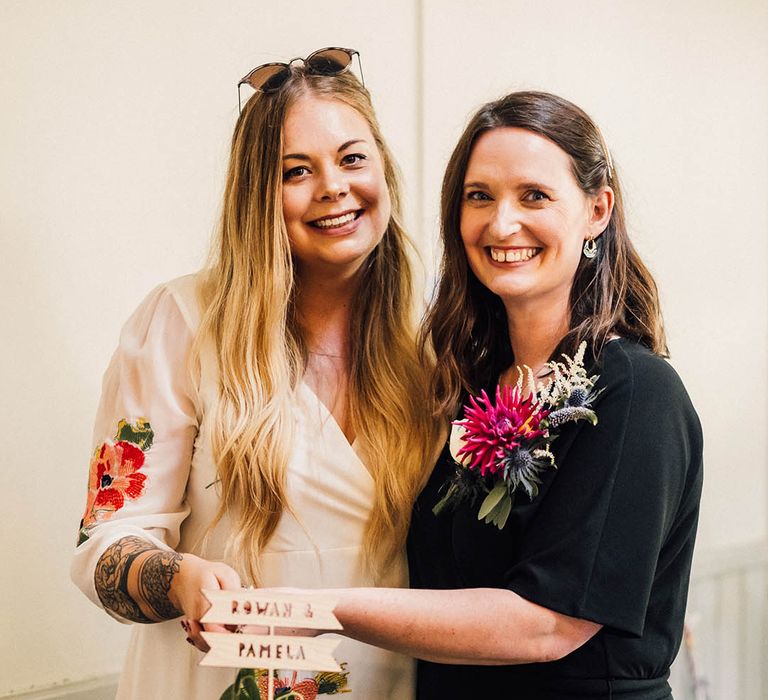 Bride in colourful embroidered wedding dress and bride in black bridal jumpsuit cut their single tier wildflower pressed wedding cake 