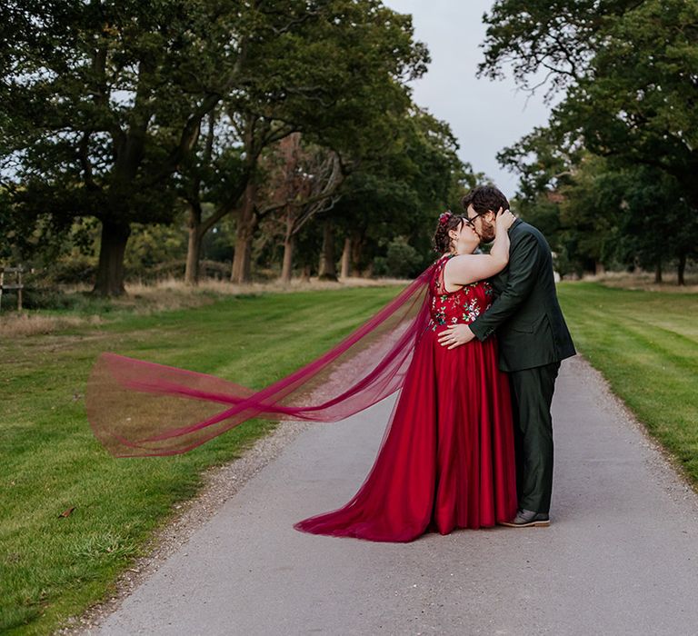 Groom in green wedding suit posing with the bride in a red wedding dress with floral embroidered details 