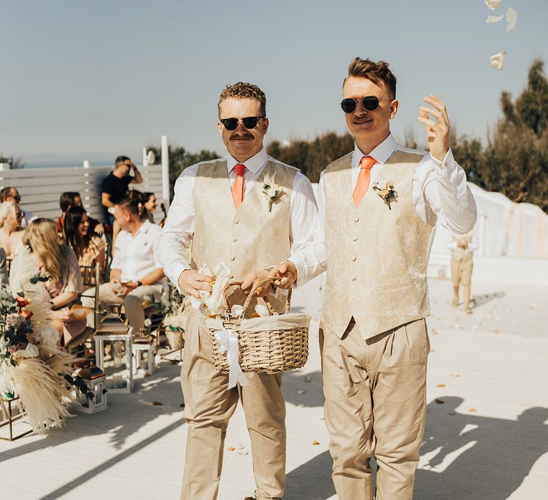 Flower men walking down the aisle together in matching suits scattering petals at their Santorini wedding 