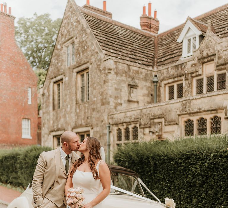 The bride and groom kiss next to the classic white vintage wedding car transport decorated with white ribbon 