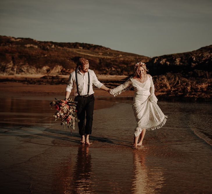 Smiling bride and groom walk along the beach in a boho and relaxed style of dress 