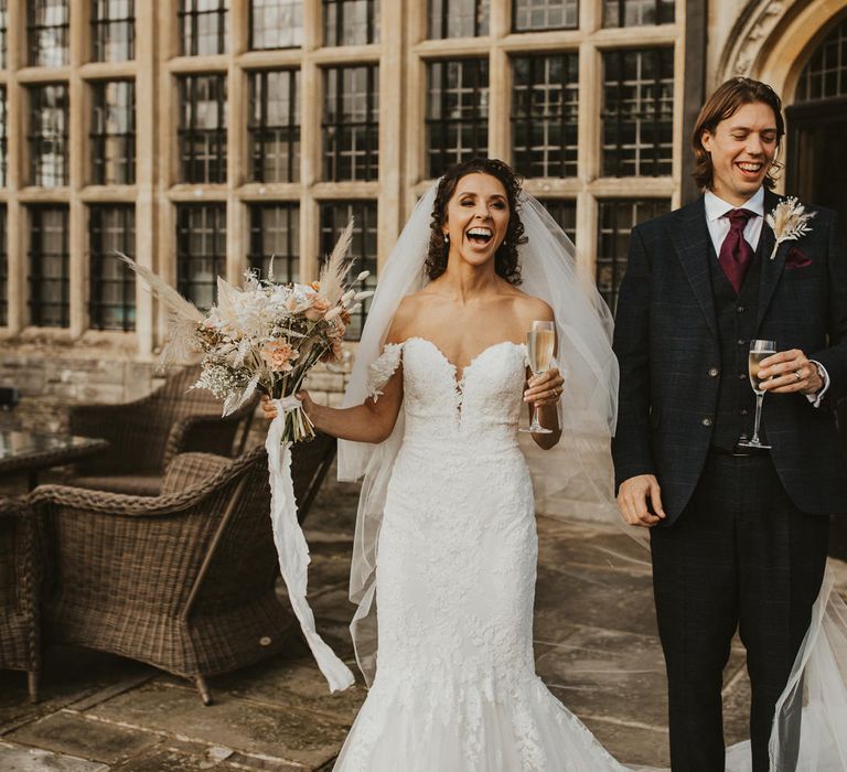 Bride with brown curly hair in a strapless lace wedding dress laughing and sipping champagne with her groom in a navy check suit