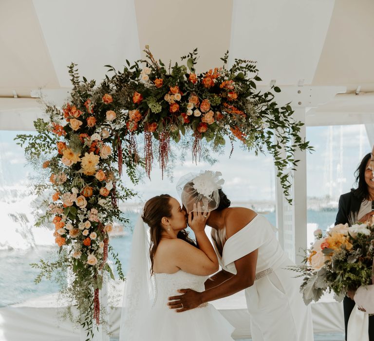 Black bride in a wedding jumpsuit and blush veil kissing her bride in a princess wedding dress at the altar 