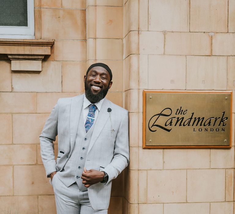 Black groom in light grey suit with blue paisley tie standing outside The Landmark London wedding venue 